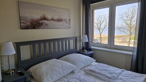 A bedroom with a bed, a nightstand, a lamp, a window with a view of the outdoors, and a painting on the wall. - Bernsteinland Barth (Barth)