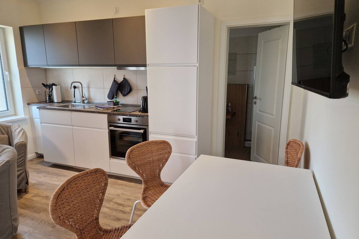 A modern kitchen with white cabinets, a built-in oven, and a dining area with wicker chairs.