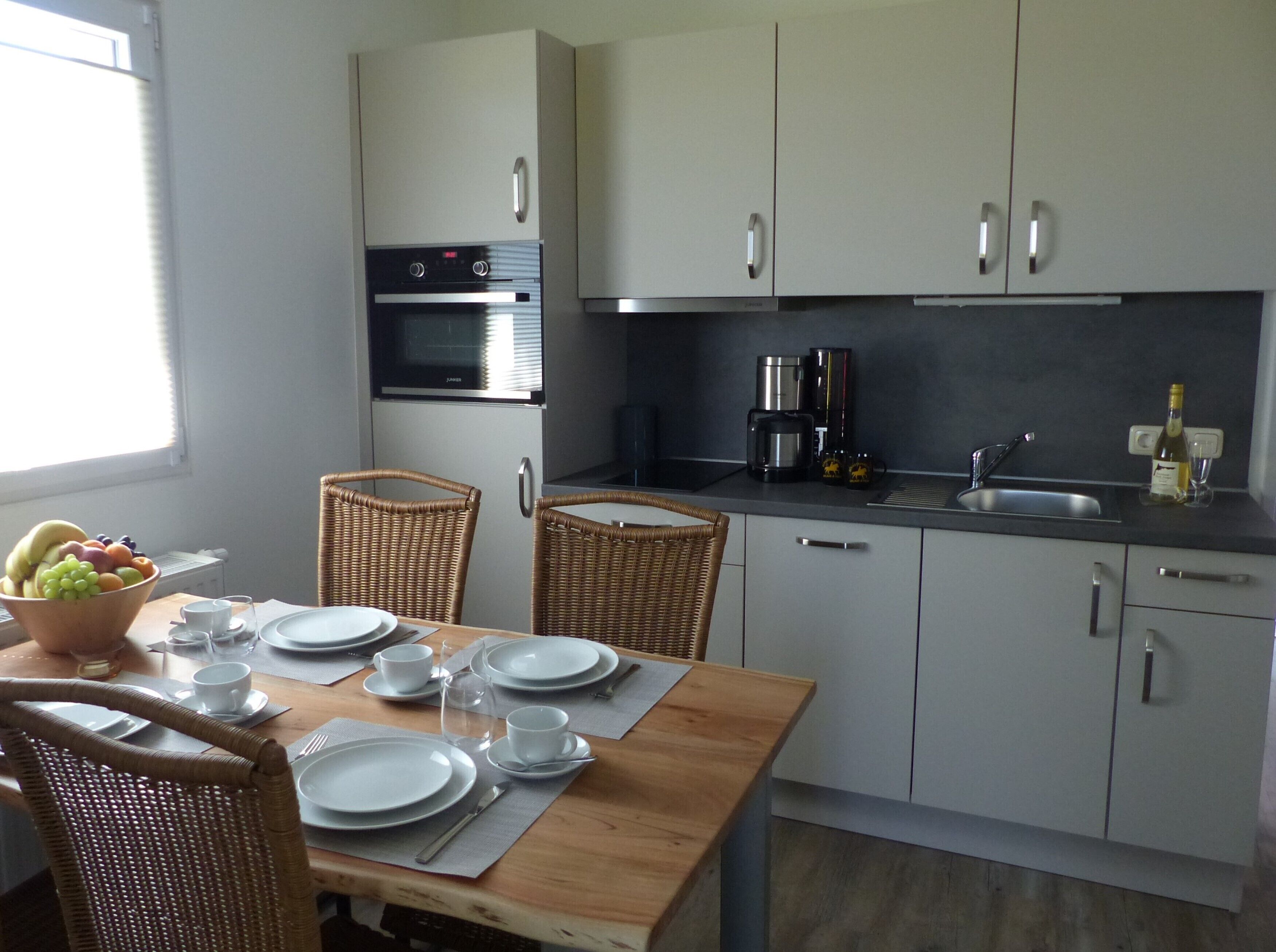 A modern kitchen with white cabinets, a dining table set for four, wicker chairs, and a fruit bowl.