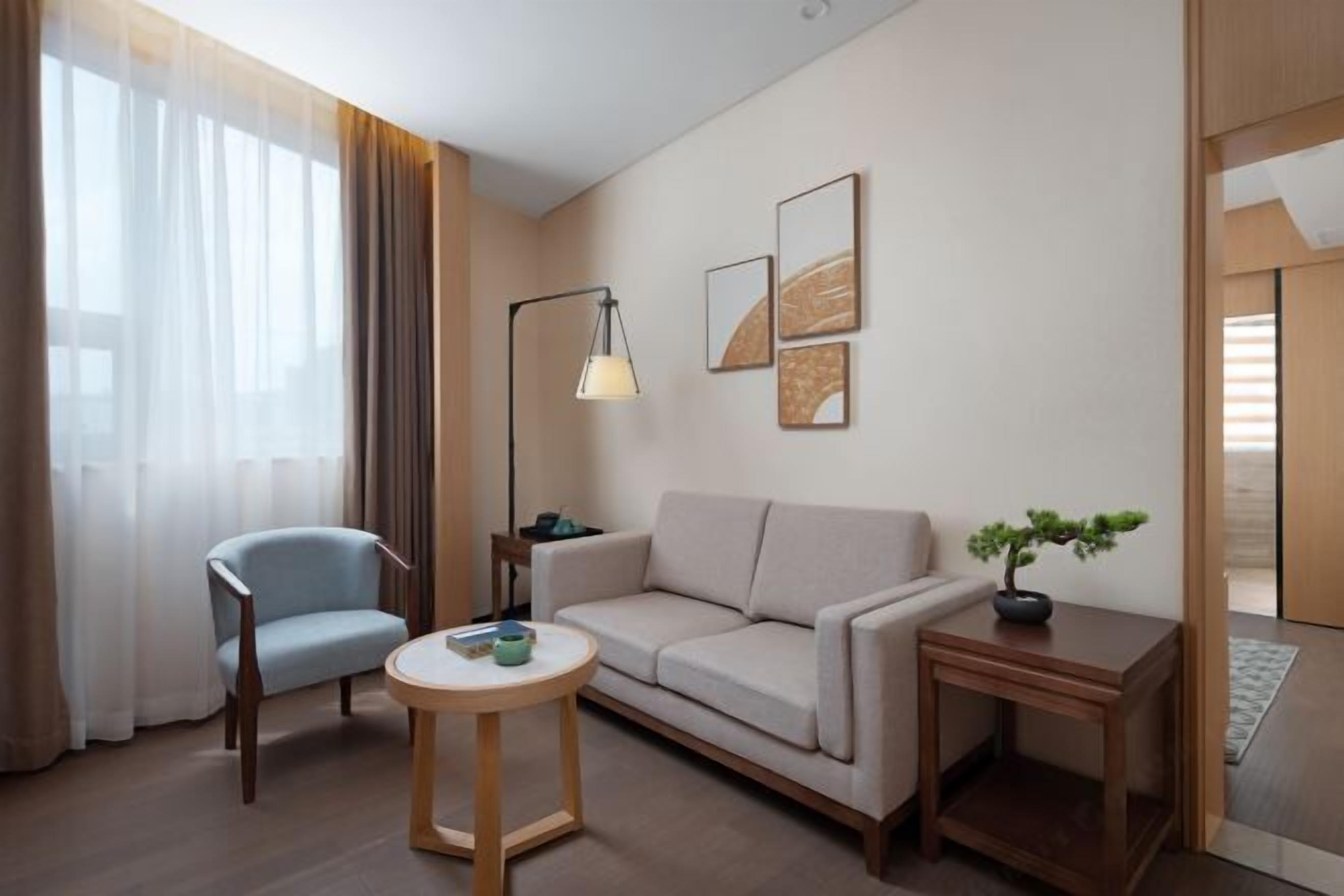 Una habitación de hotel moderna con sofá, sillón, mesa redonda, lámpara de pie y una obra de arte en la pared.