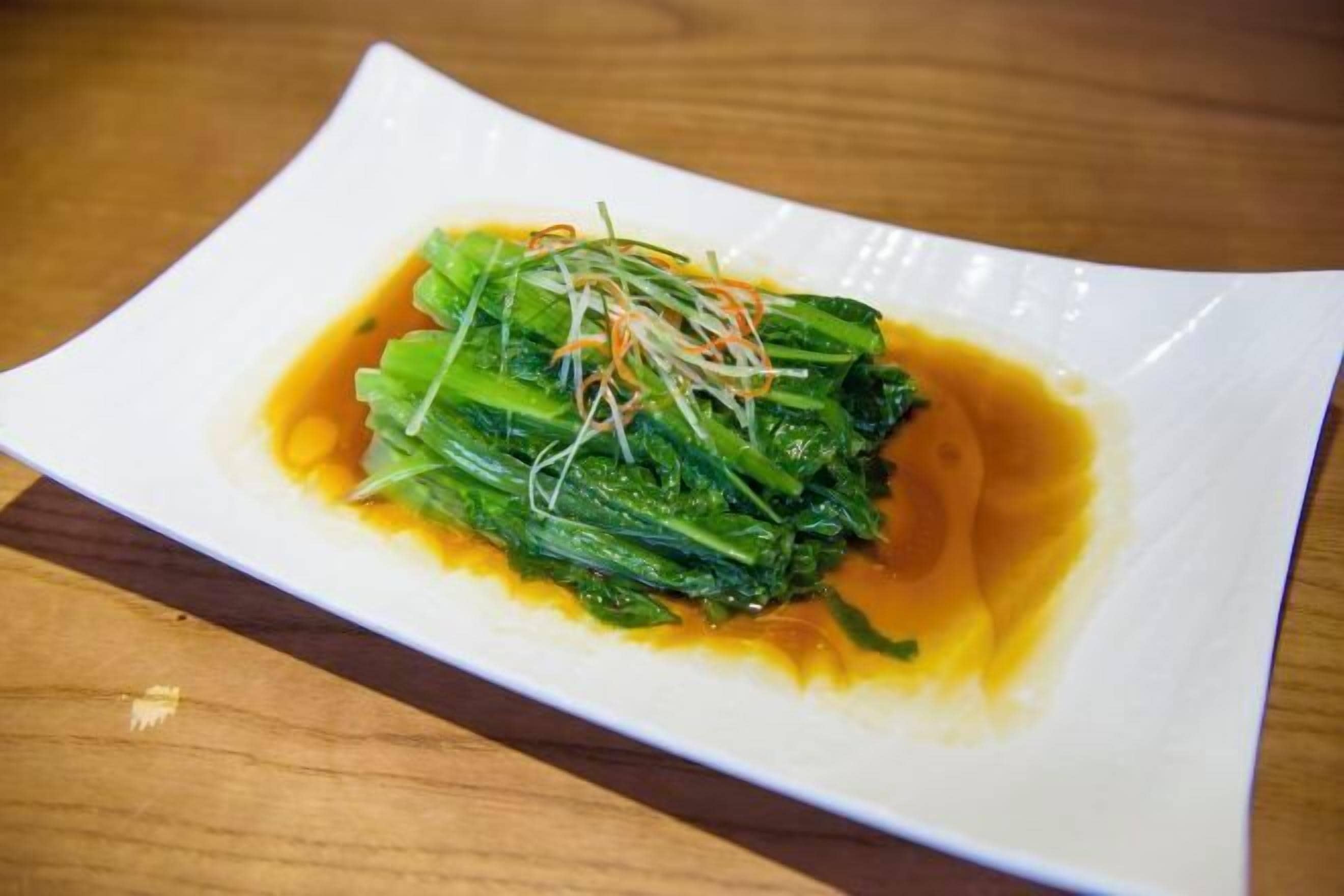 A white plate with a dish of green leafy vegetables in a light brown sauce, placed on a wooden table.