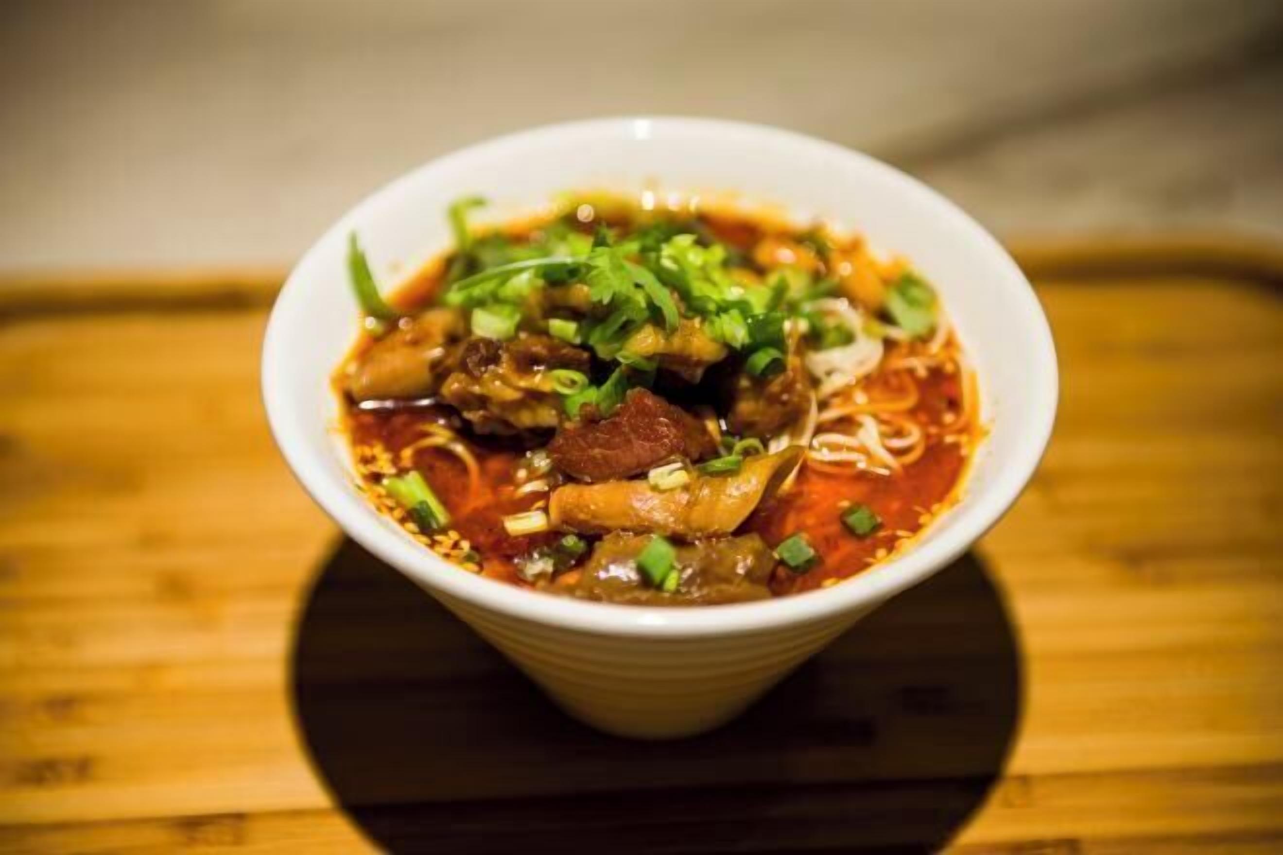 A bowl of soup with noodles and meat, garnished with green herbs, on a wooden surface.