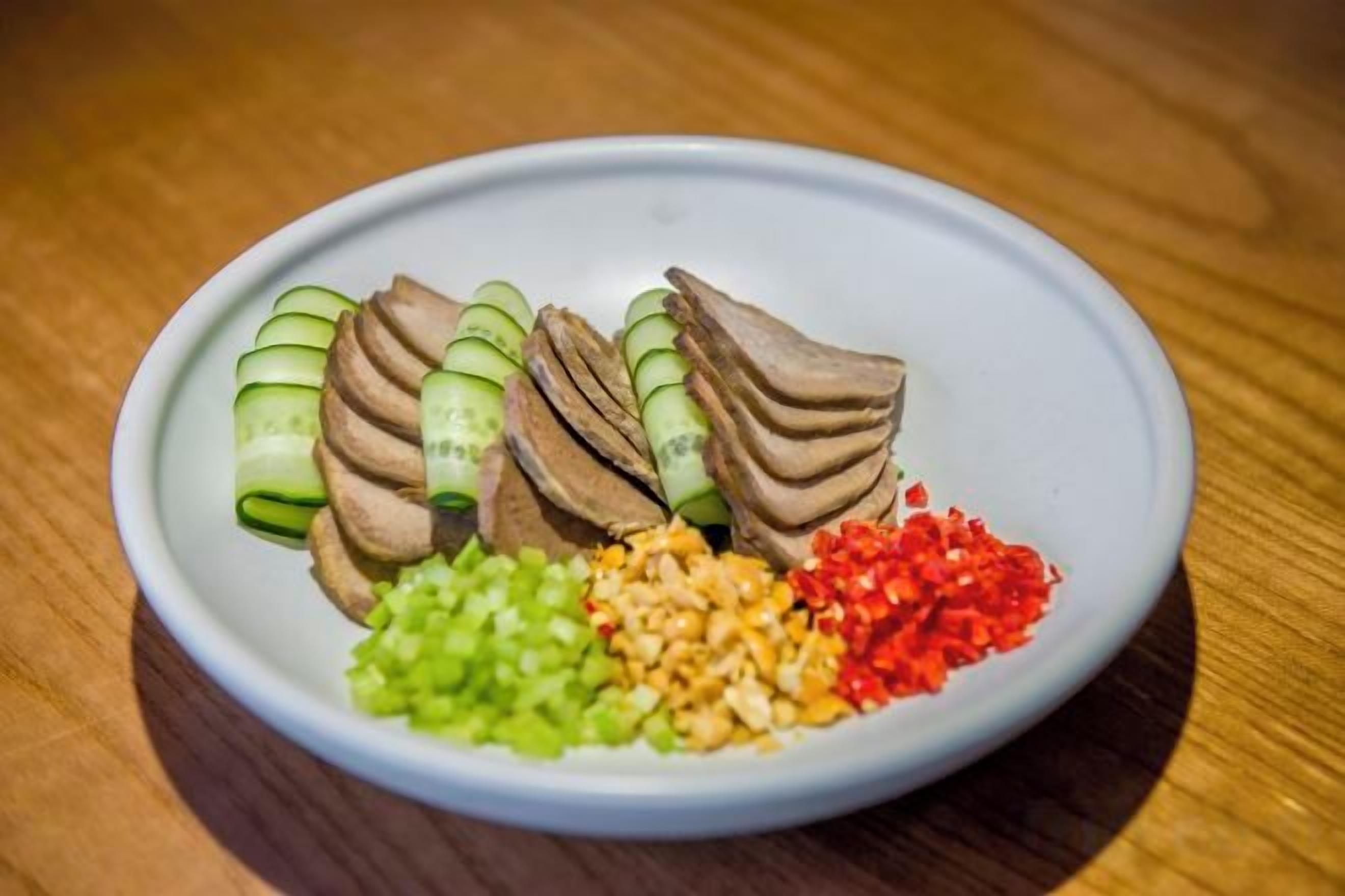 Une assiette contenant des tranches de concombre, un aliment de couleur brunâtre, ainsi que des légumes verts et rouges coupés en morceaux.