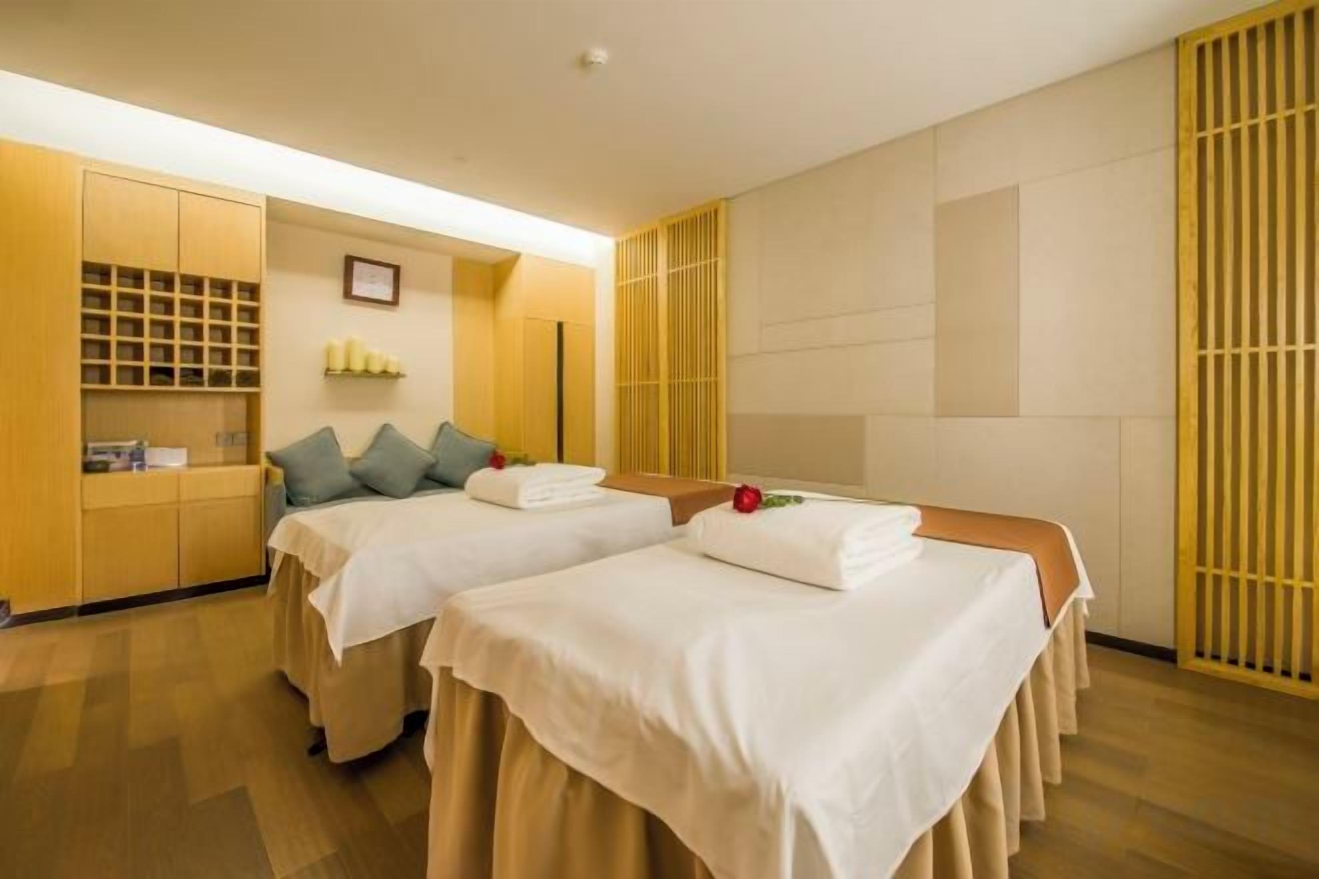 A hotel room with two beds, a wooden wardrobe, a small table, and a wall-mounted shelf.