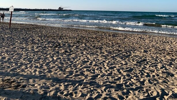 Ein sandiger Strand mit Fußspuren, die zum Wasser führen, ein Pier in der Ferne und ein klarer, blauer Himmel.