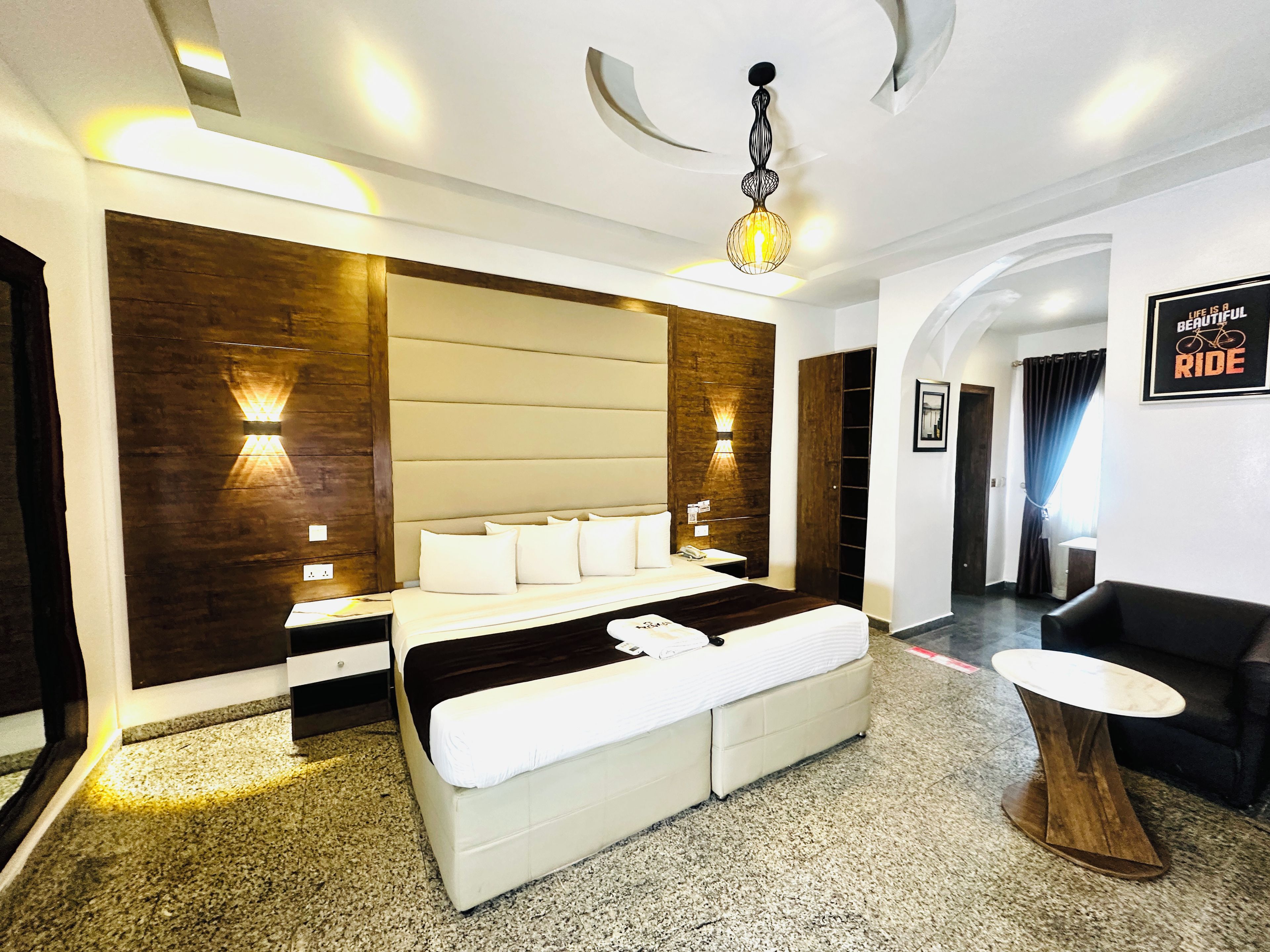 A modern hotel room with a large bed, a wooden accent wall, a small seating area, and a ceiling light fixture.
