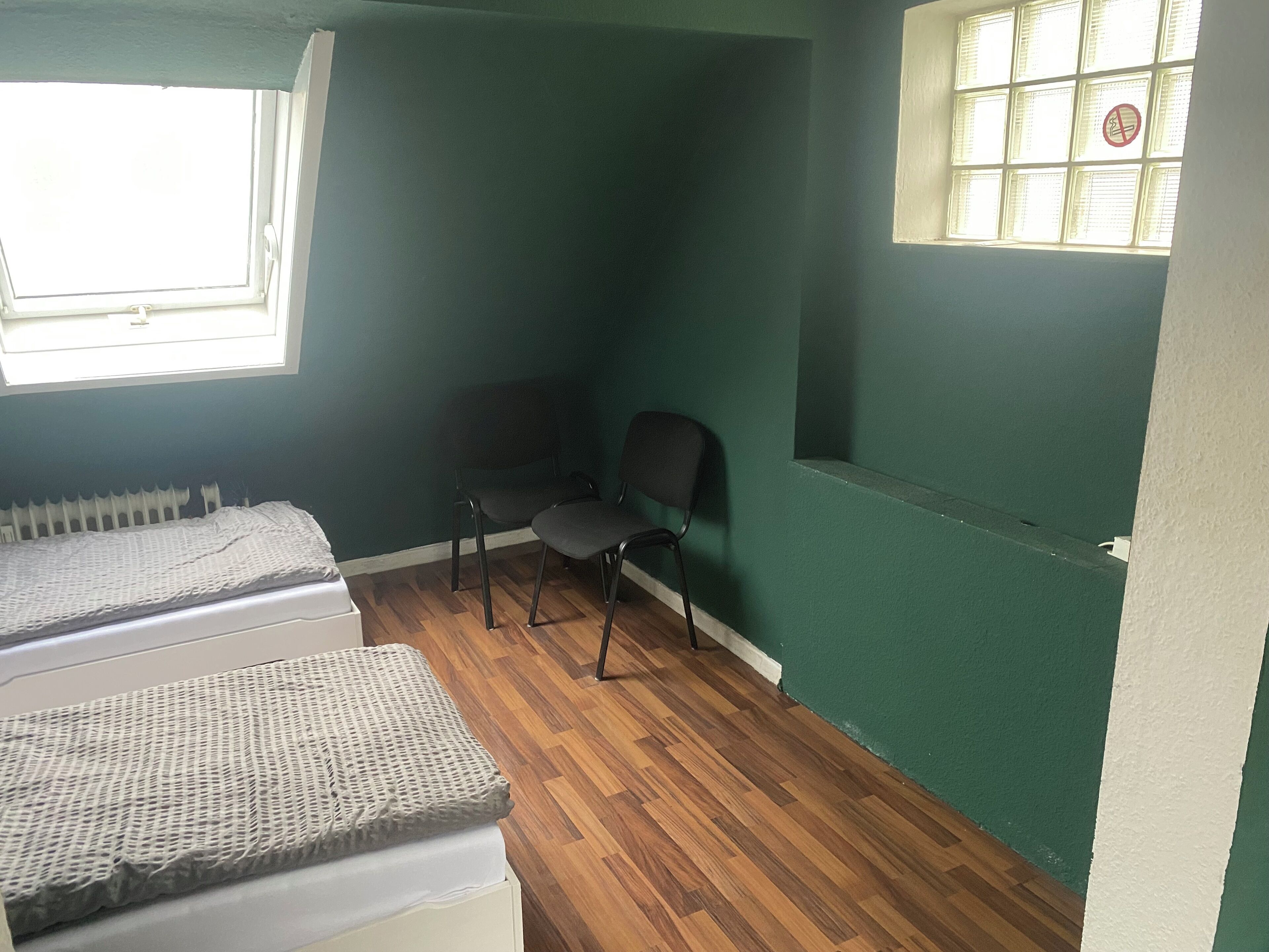 Ein Zimmer mit zwei Betten, einem Sessel, einem Heizkörper und einem Fenster mit Jalousien.