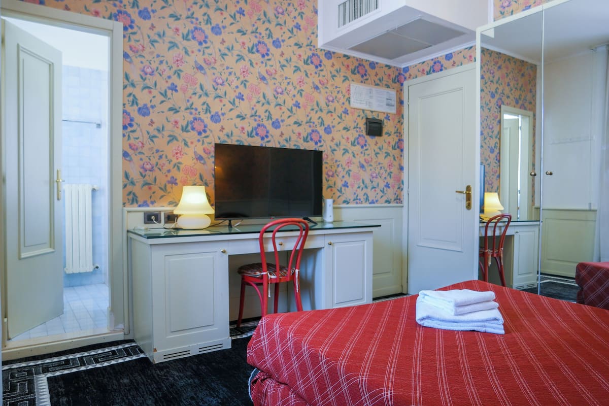 A hotel room with a bed, a desk with a computer and lamp, a chair, and a TV.