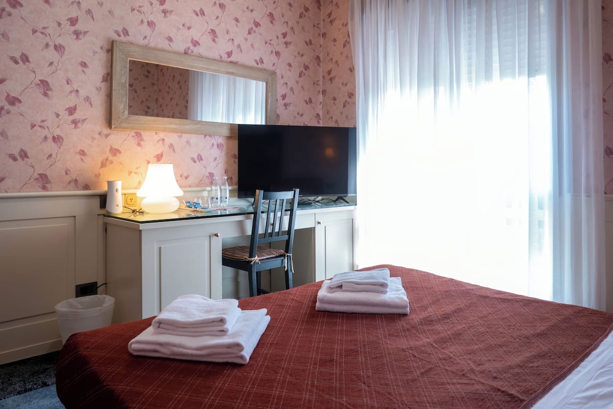A hotel room with a bed, a desk with a lamp, a chair, a TV, and a window with curtains.