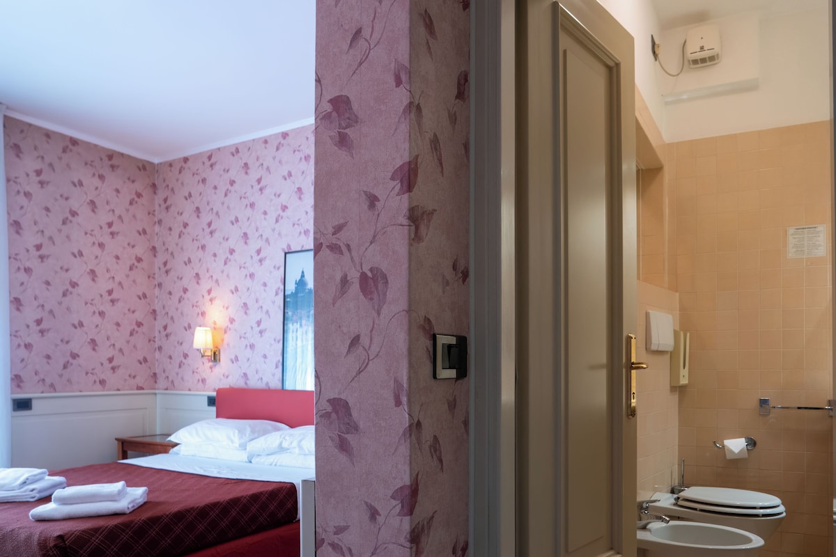 A hotel room with a bed, a nightstand, a wall-mounted lamp, and a window with a view.