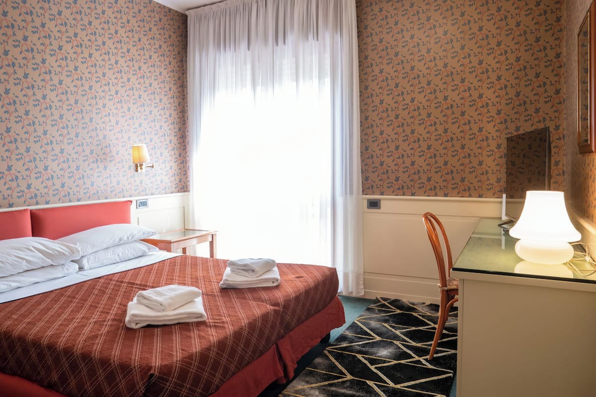 A hotel room with a bed, a desk, a chair, a lamp, and a window with curtains.