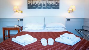 Une chambre d’hôtel avec un grand lit, deux lampes de chevet, un tableau représentant un paysage urbain, un téléphone sur une table d’appoint, ainsi que des serviettes et des chaussons sur le lit.
