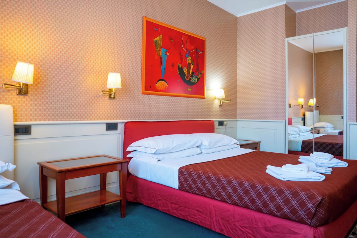 A hotel room with a red bed, white pillows, a wooden nightstand, a small table, and a large mirror.