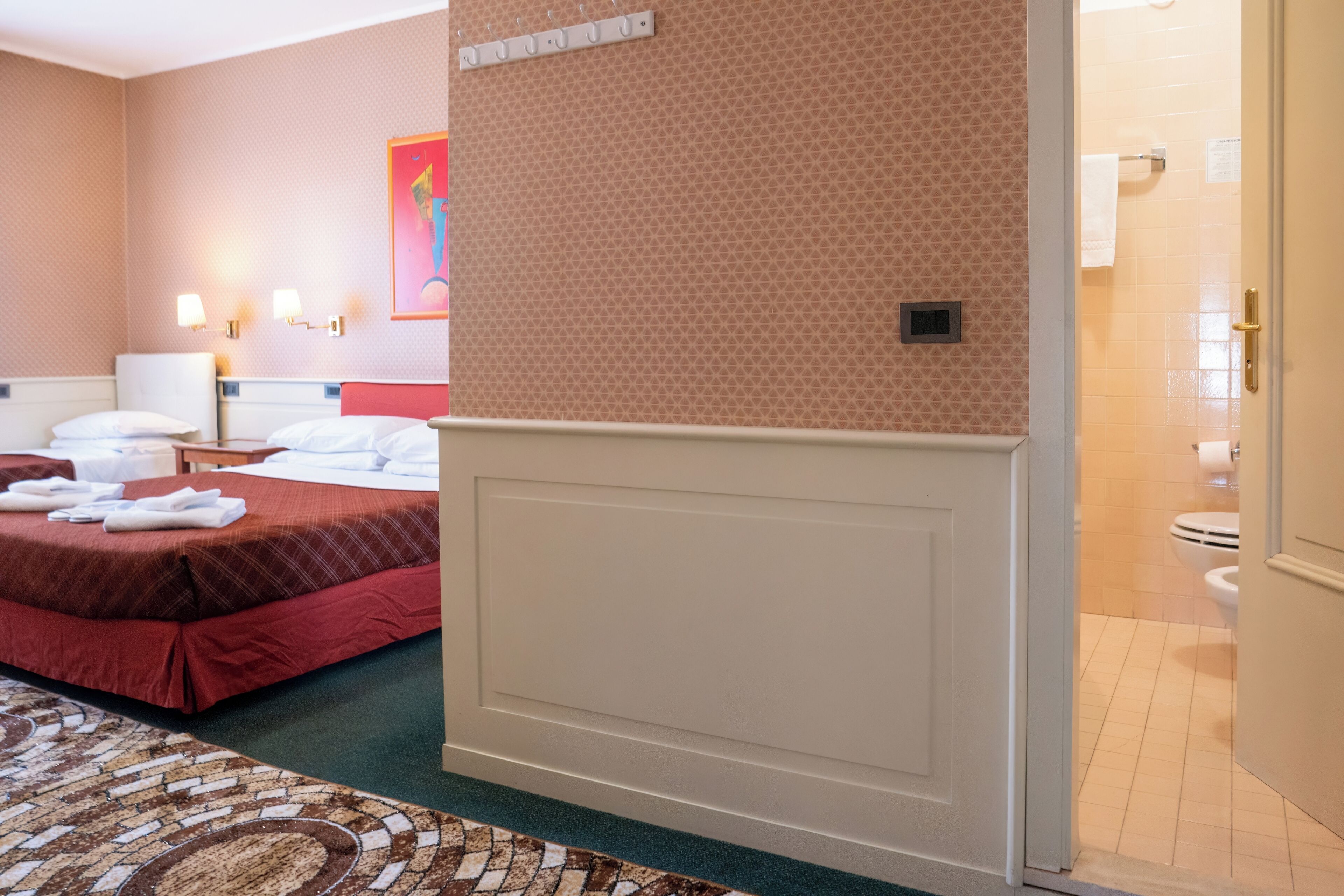 A hotel room with a bed, a nightstand, a wall-mounted lamp, and a bathroom visible through an open door.