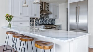 Uma cozinha moderna com armários brancos, uma ilha central com banquetas, eletrodomésticos de aço inoxidável e iluminação pendente.