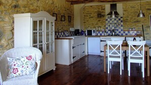 Une cuisine avec des murs en pierre, des poutres en bois, un meuble blanc, une table à manger avec des chaises et un luminaire suspendu.