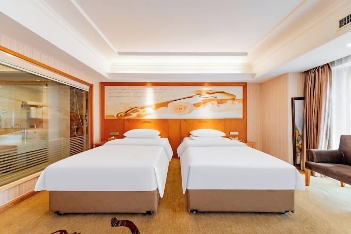 Une chambre d’hôtel avec deux lits simples, une grande fenêtre avec stores, une œuvre d’art accrochée au mur et un fauteuil en cuir marron.