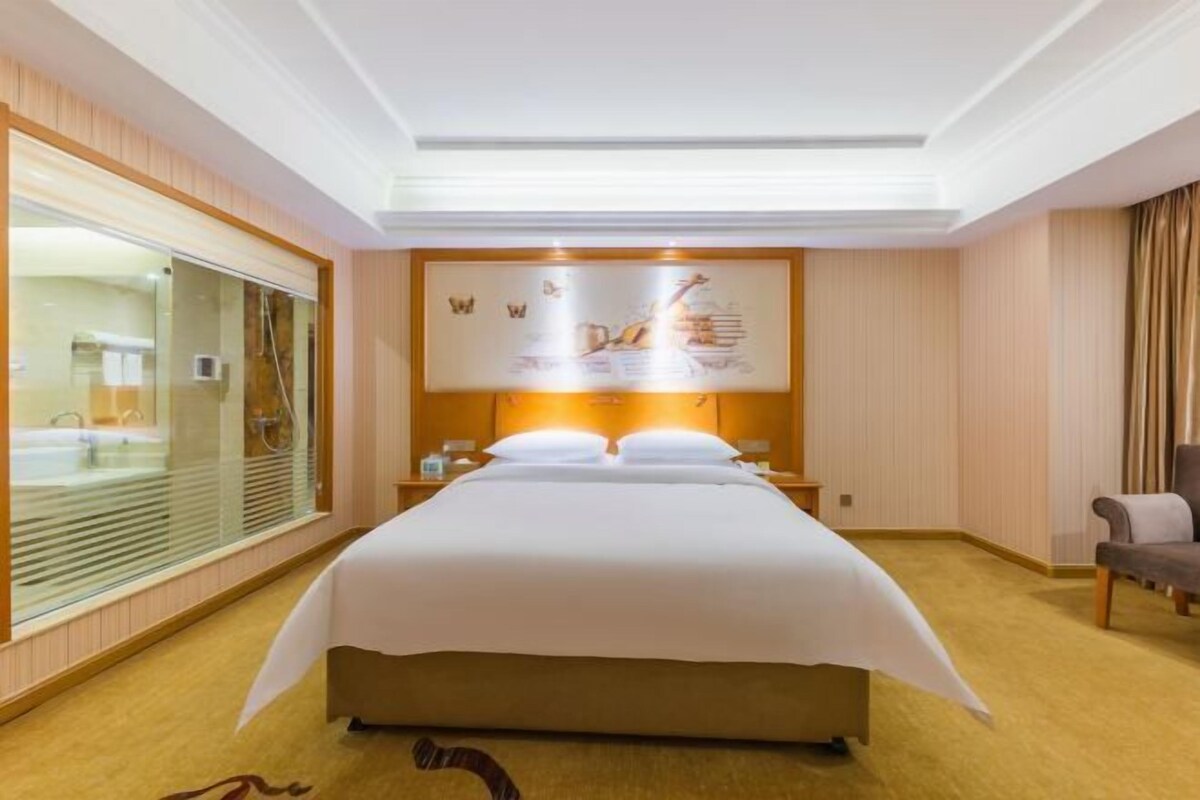 A hotel room with a large bed, a glass-enclosed shower area, a wooden floor, and a wall-mounted artwork.