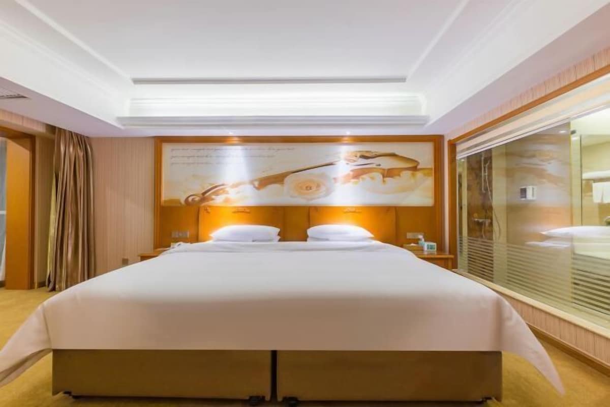 Une chambre d’hôtel avec un grand lit, une tête de lit ornée d’un panneau décoratif, une grande fenêtre avec des stores et un miroir sur le mur.