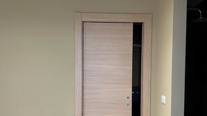 Una habitación con puerta de madera, un banco blanco, un otoman redondo y una toalla sobre una encimera.