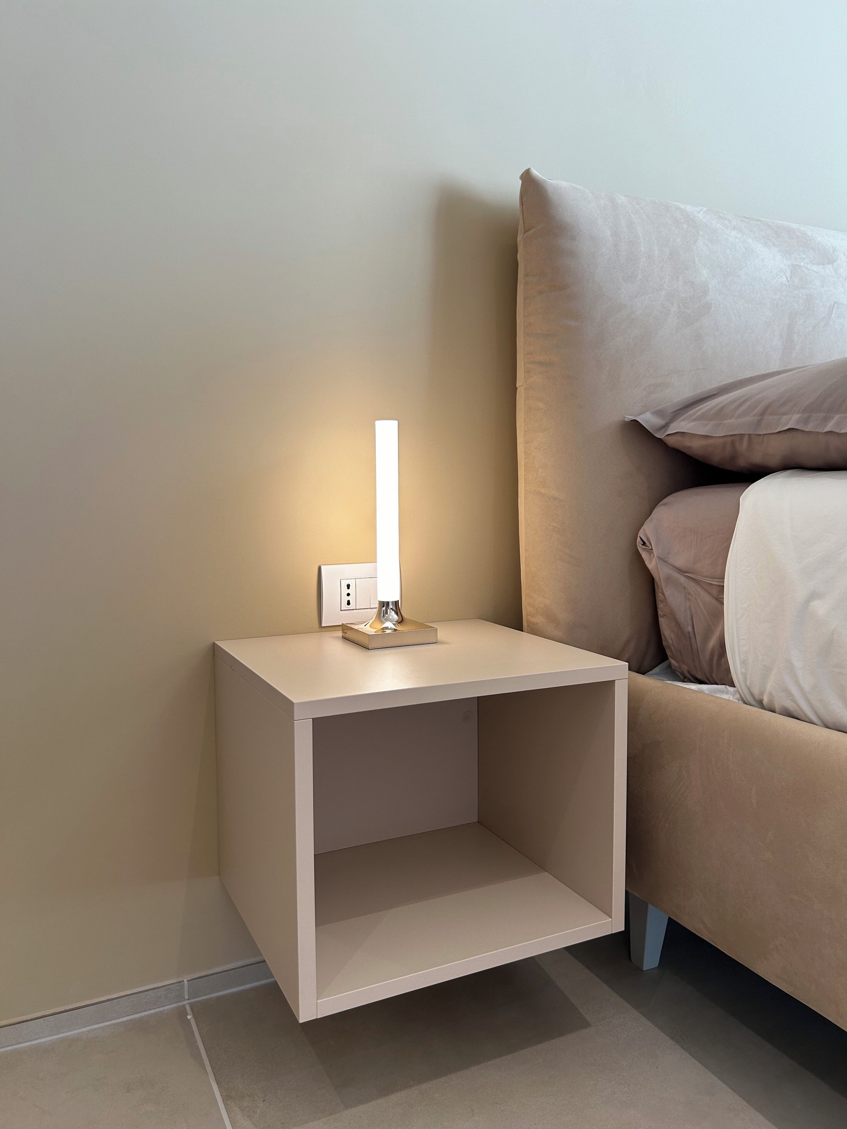 A minimalist bedside table with a lamp, a bed with beige bedding, and a plain wall.