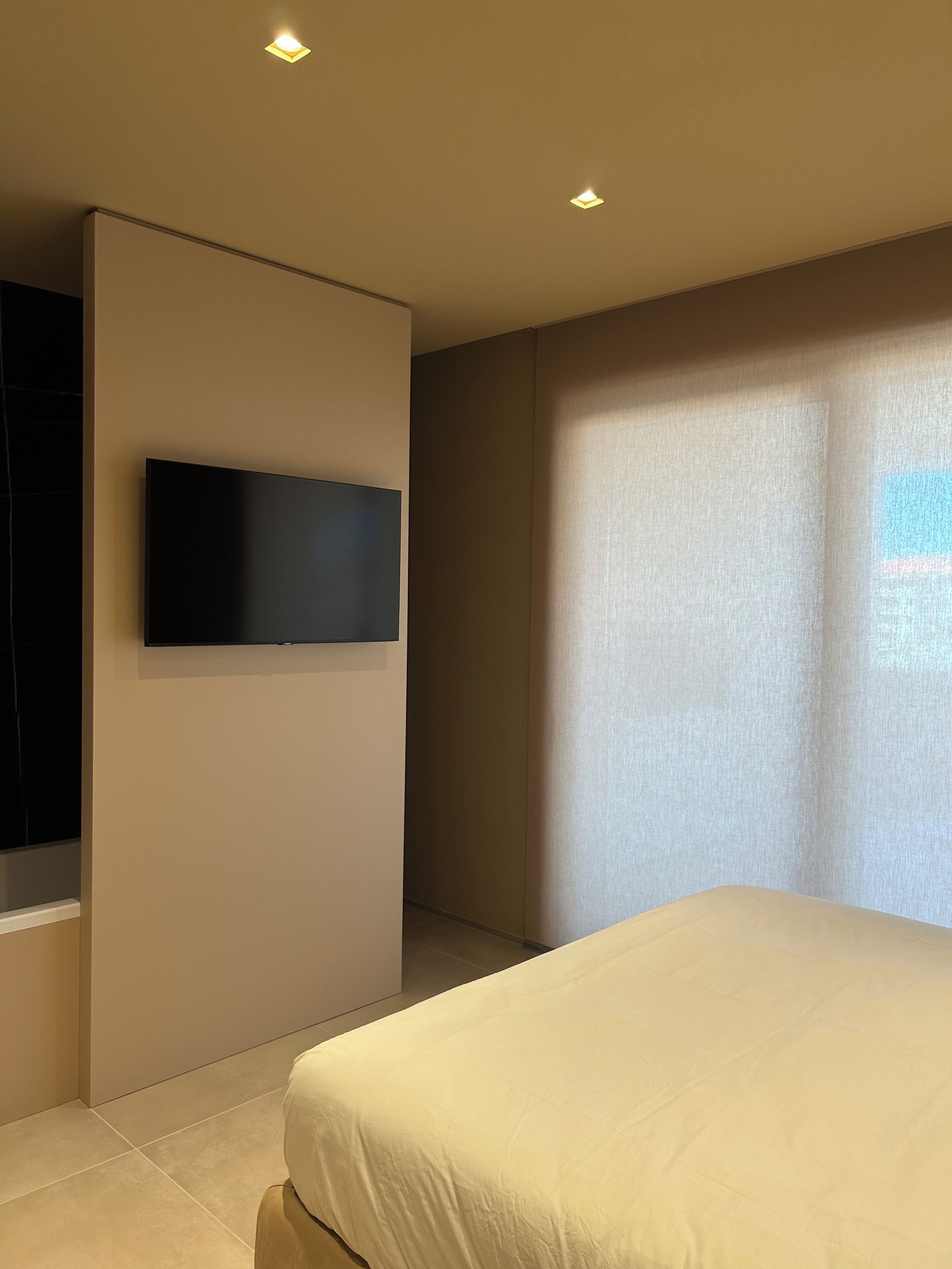 A modern hotel room with a large bed, a flat-screen TV mounted on the wall, and a large window providing natural light.