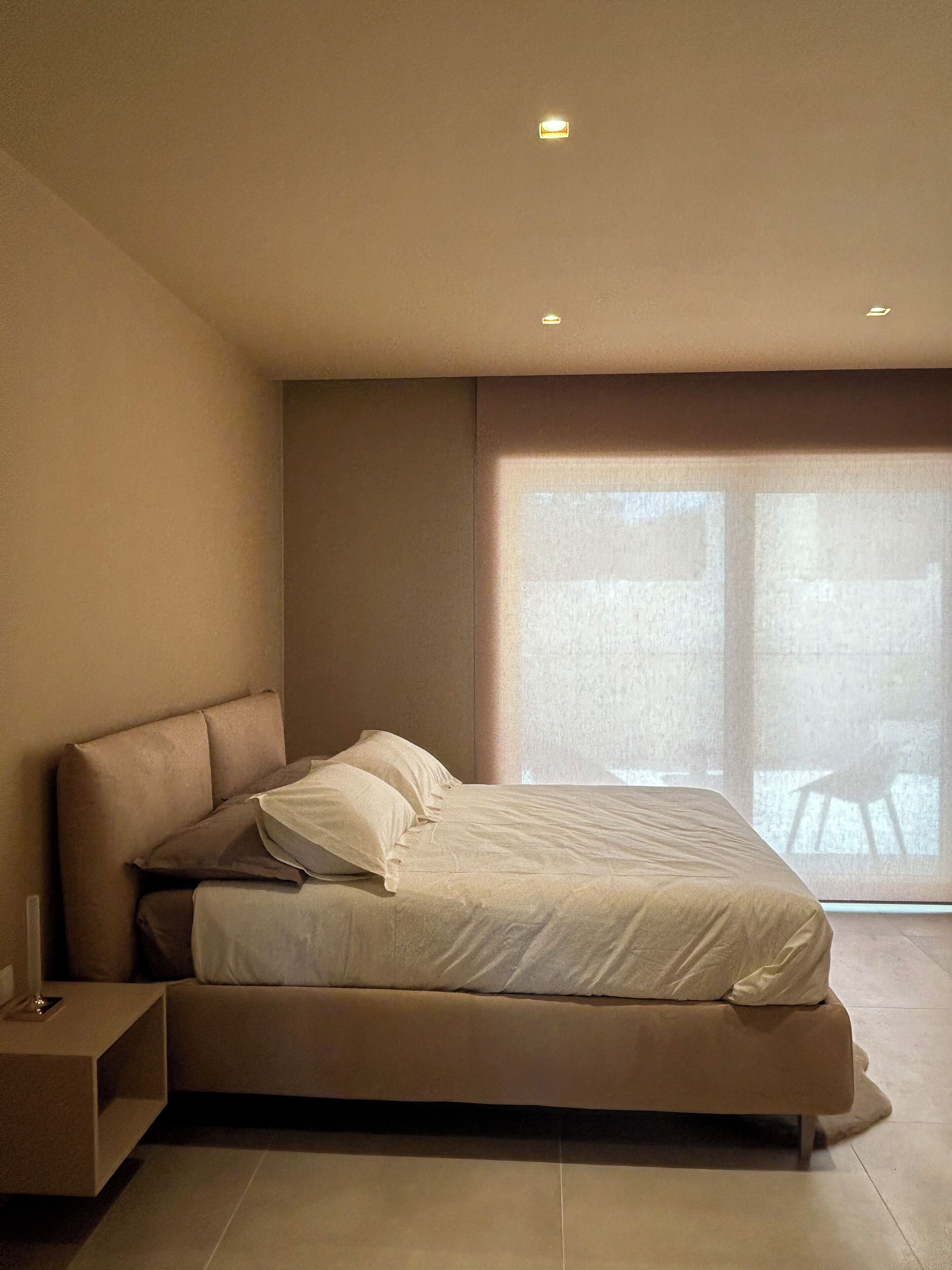 A modern bedroom with a large bed, a nightstand, and a window with a view.