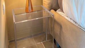 Una mesita de noche de cristal transparente con una lámpara, un sofá color beige y una cama con ropa de cama blanca.