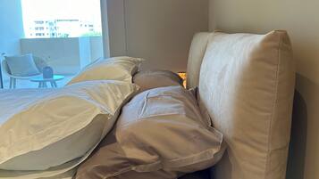 A bed with white and beige bedding, a chair, and a small table with a lamp.