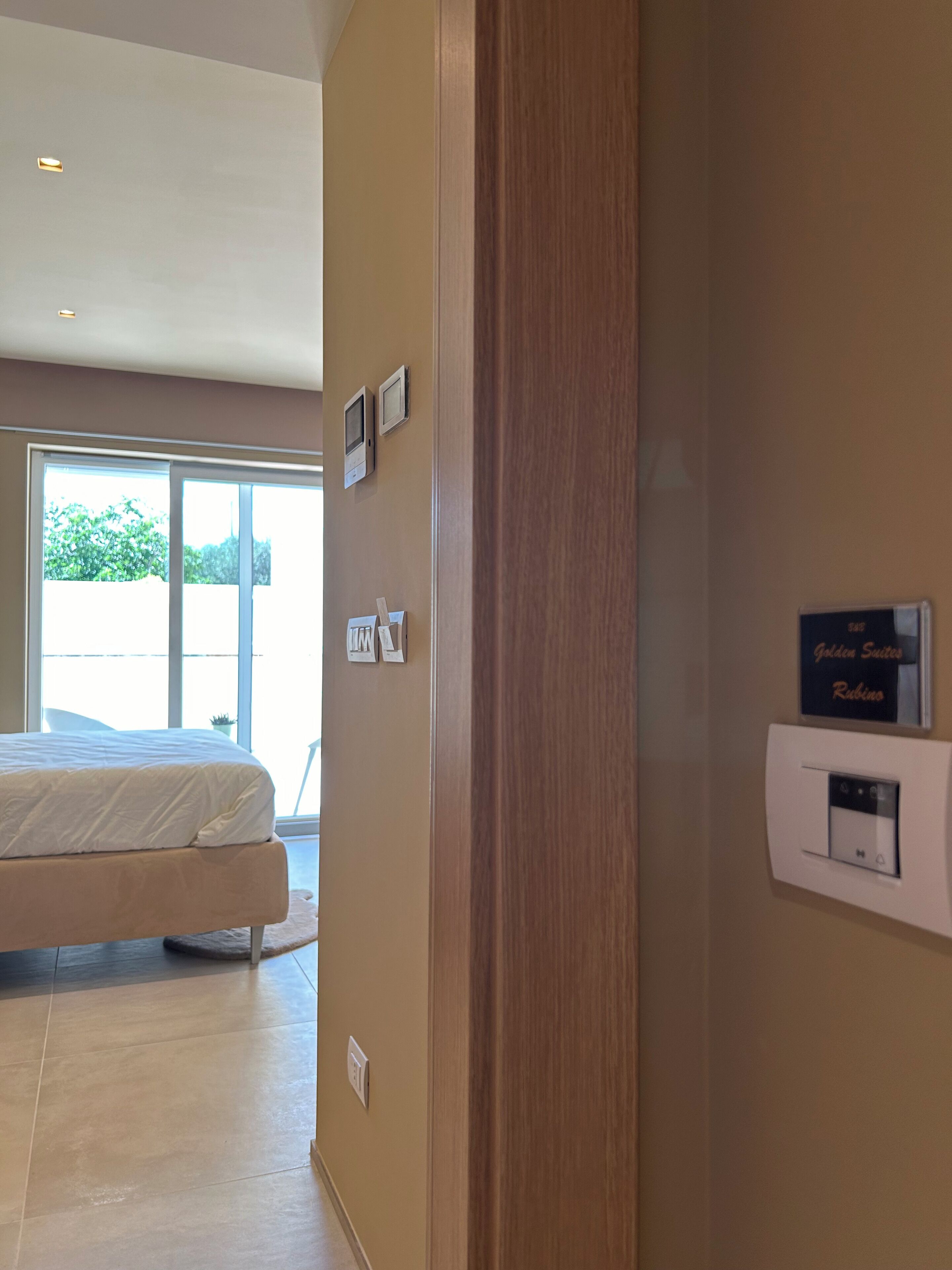 A hotel room with a bed, a large window, a wall-mounted thermostat, and a door.