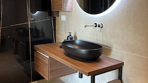 Un baño moderno con tocador de madera, un lavamanos tipo cuenco negro, un espejo redondo con iluminación integrada y un perchero para toallas.
