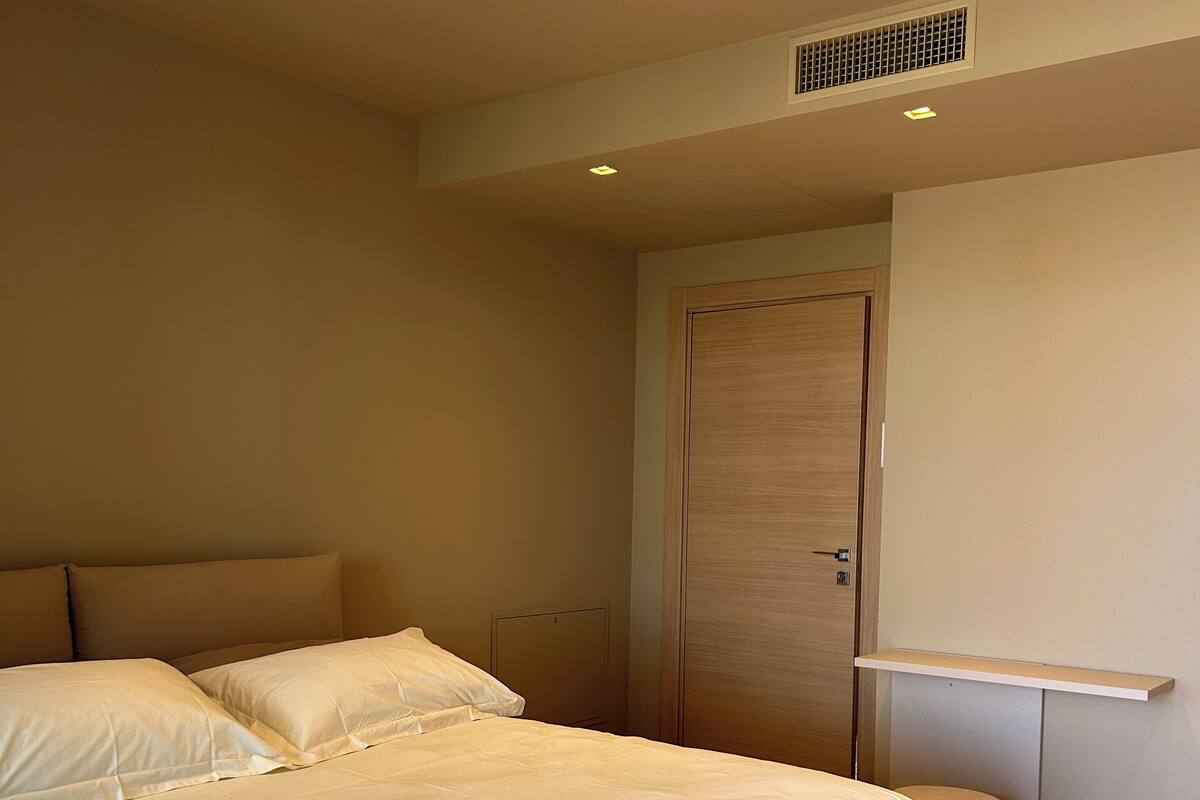 Une chambre moderne avec deux lits, une armoire en bois, un petit pouf rond et des spots encastrés au plafond.