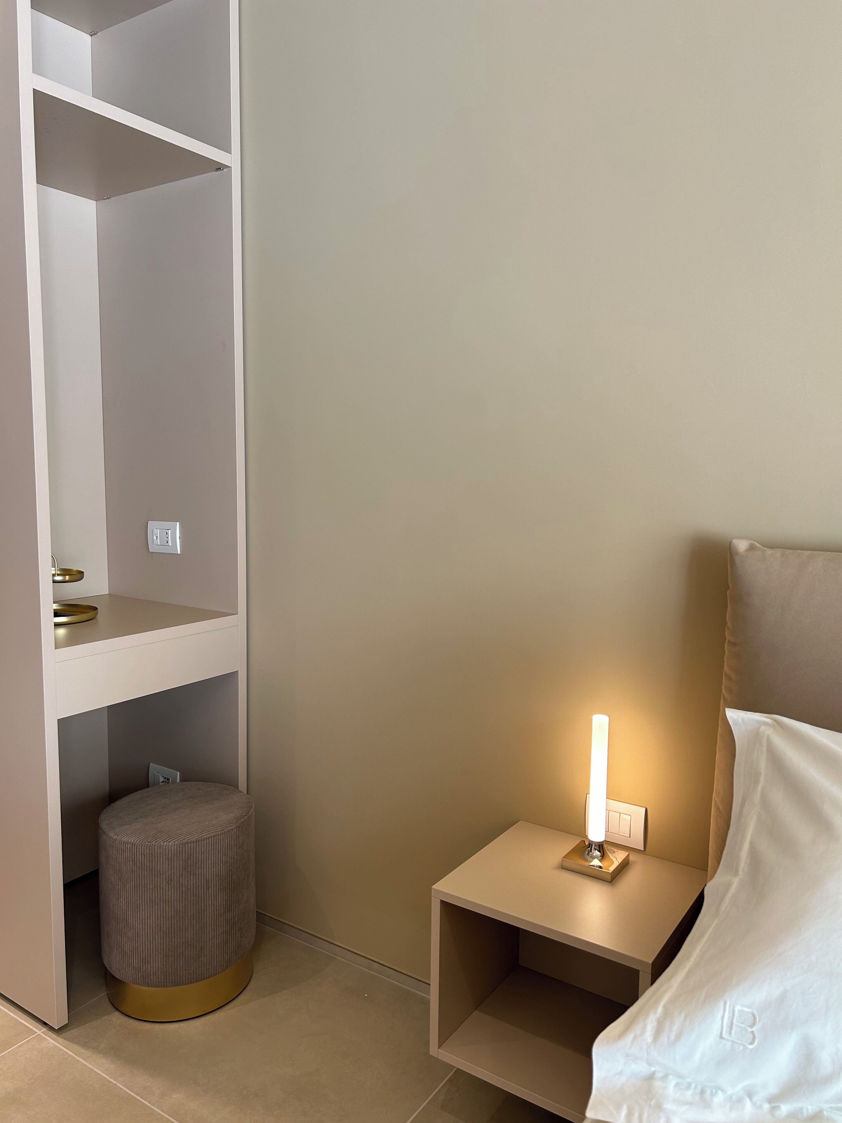A bedroom with a bed, bedside table with a lamp, a small stool, and a built-in shelving unit.