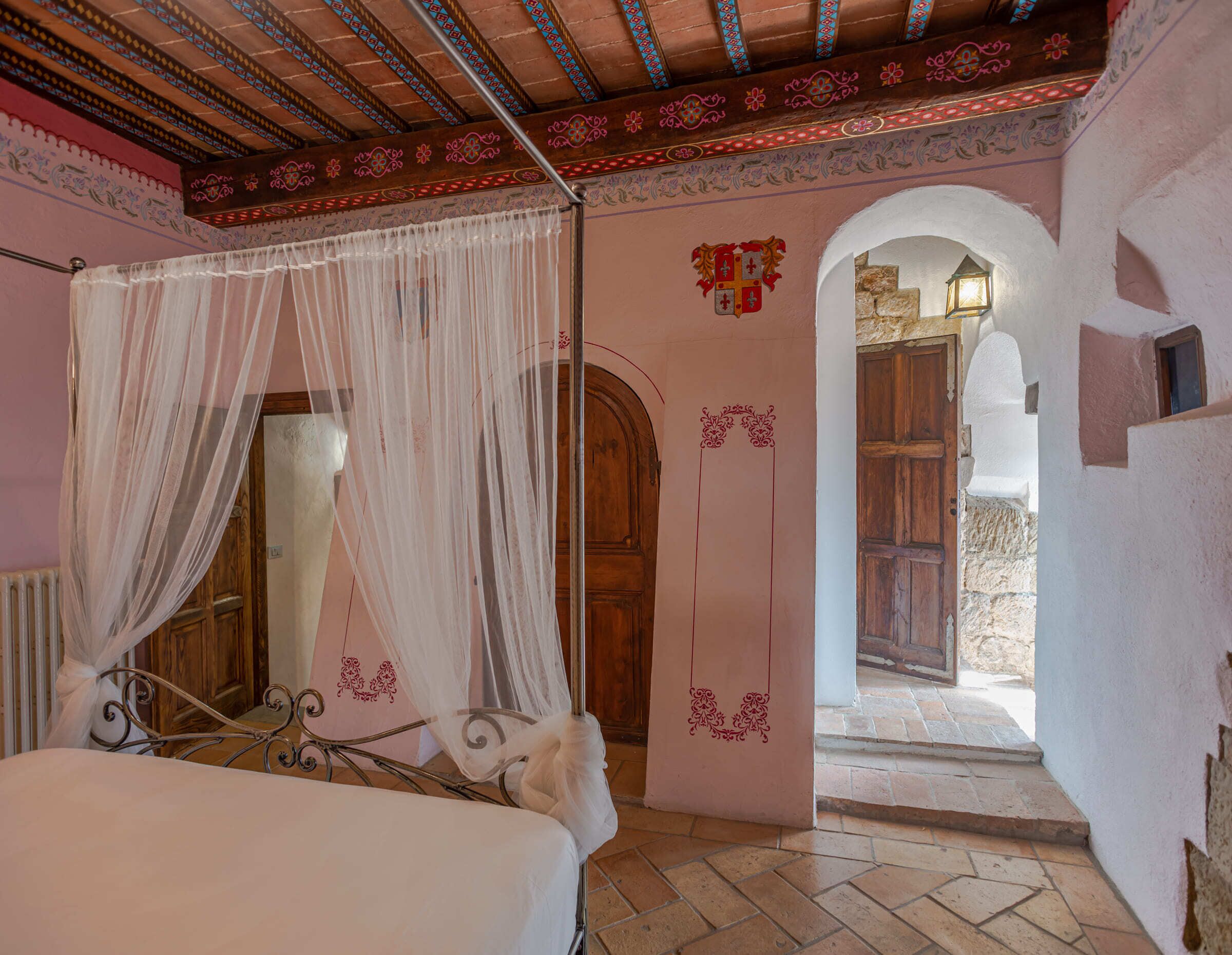 Un dormitorio con una cama, una puerta de madera, un escudo decorativo y un camino de piedra que lleva al exterior.