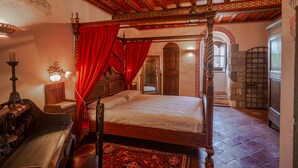 Een hemelbed met rode gordijnen, een houten bureau met een lamp en een kamerbreed tapijt met een patroon.