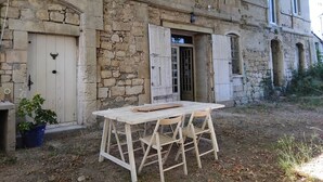 Un edificio rústico de piedra con una mesa y sillas de madera en el frente, una puerta y una pequeña planta en maceta.