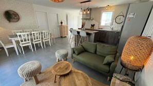 Una sala de estar moderna con un sofá verde, una mesa de centro de madera y otomanas redondas. Se ve una cocina con una mesa de comedor y sillas al fondo.