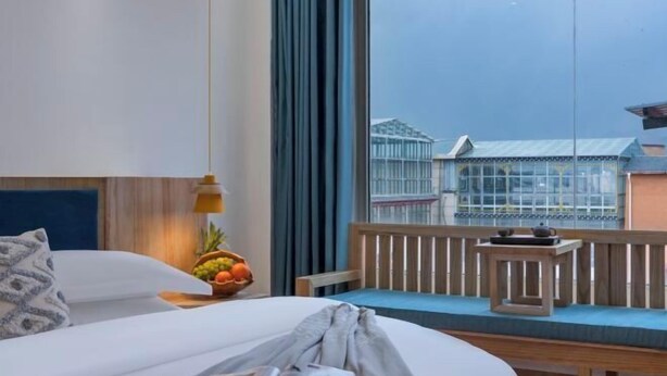 Habitación de hotel con una cama, un ventanal con vistas a edificios, una mesita con fruteros y un banco junto a la ventana.