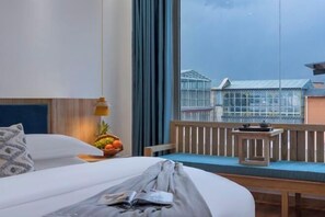 Une chambre d’hôtel avec un lit, une fenêtre donnant sur des immeubles, une petite table avec un panier de fruits et un banc près de la fenêtre.