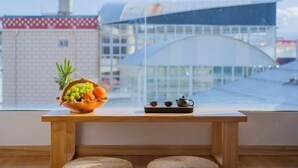 Una mesa de madera con un cuenco de fruta y una tetera, una ventana con vistas a edificios.