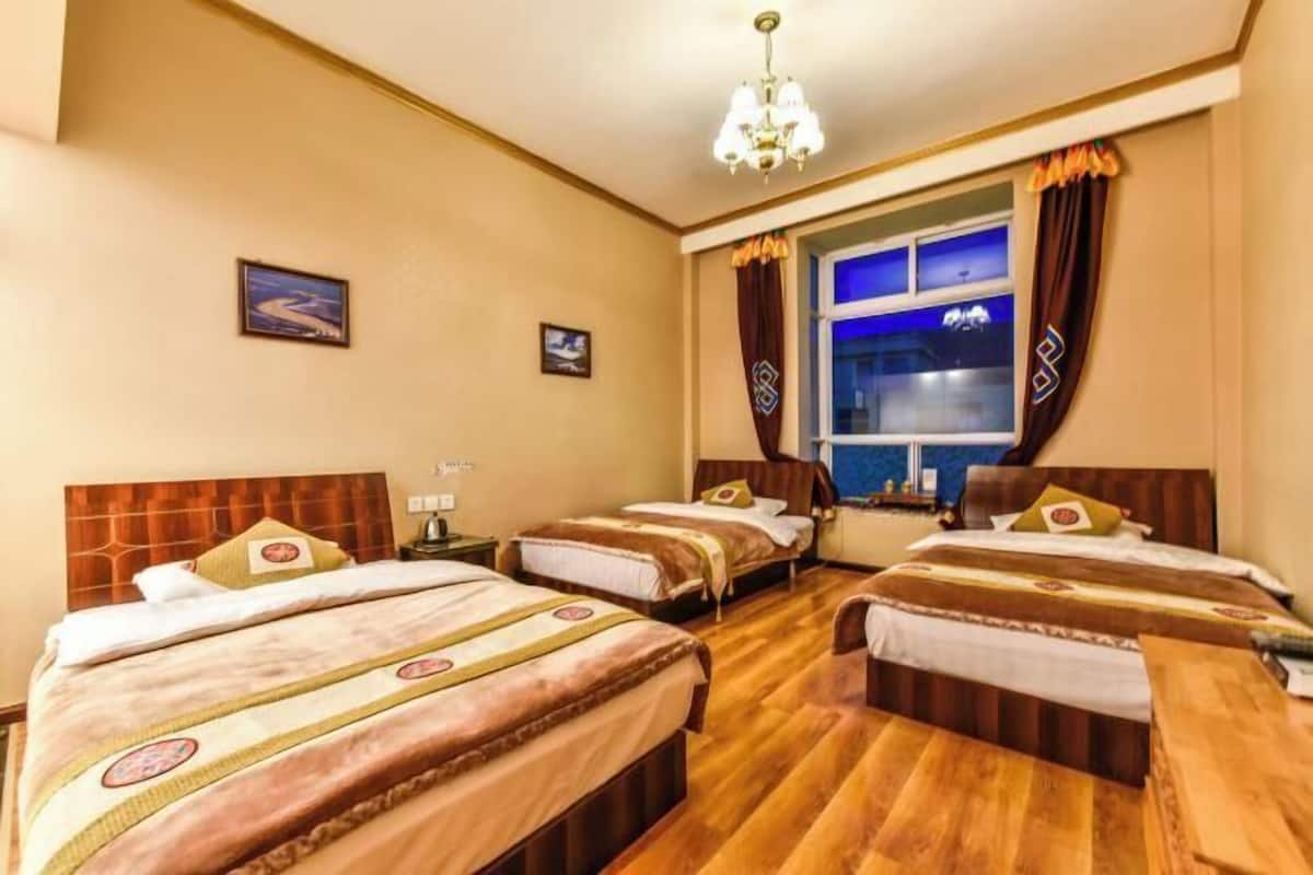 Une chambre d’hôtel avec trois lits, un lustre, une fenêtre avec des rideaux et une vue sur la ville la nuit.