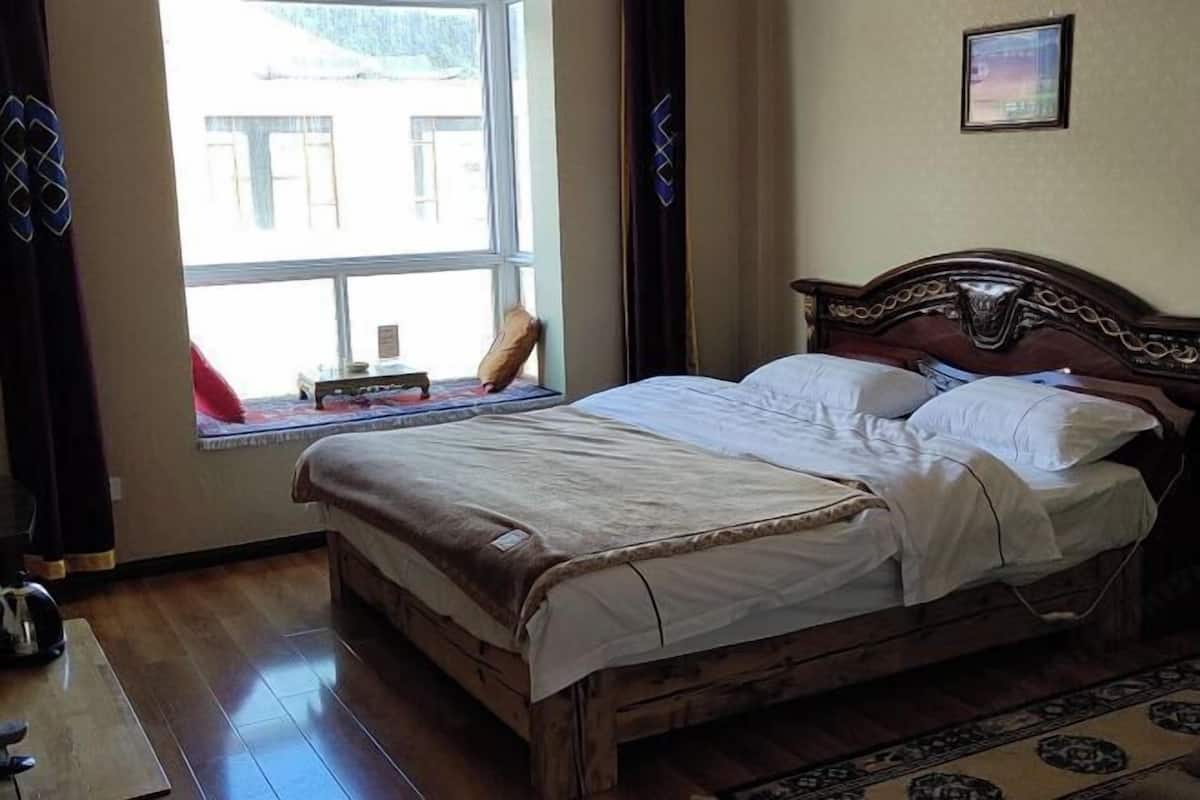 Un lit en bois recouvert de draps blancs, une fenêtre donnant sur des immeubles et une petite table avec un livre et une lampe.