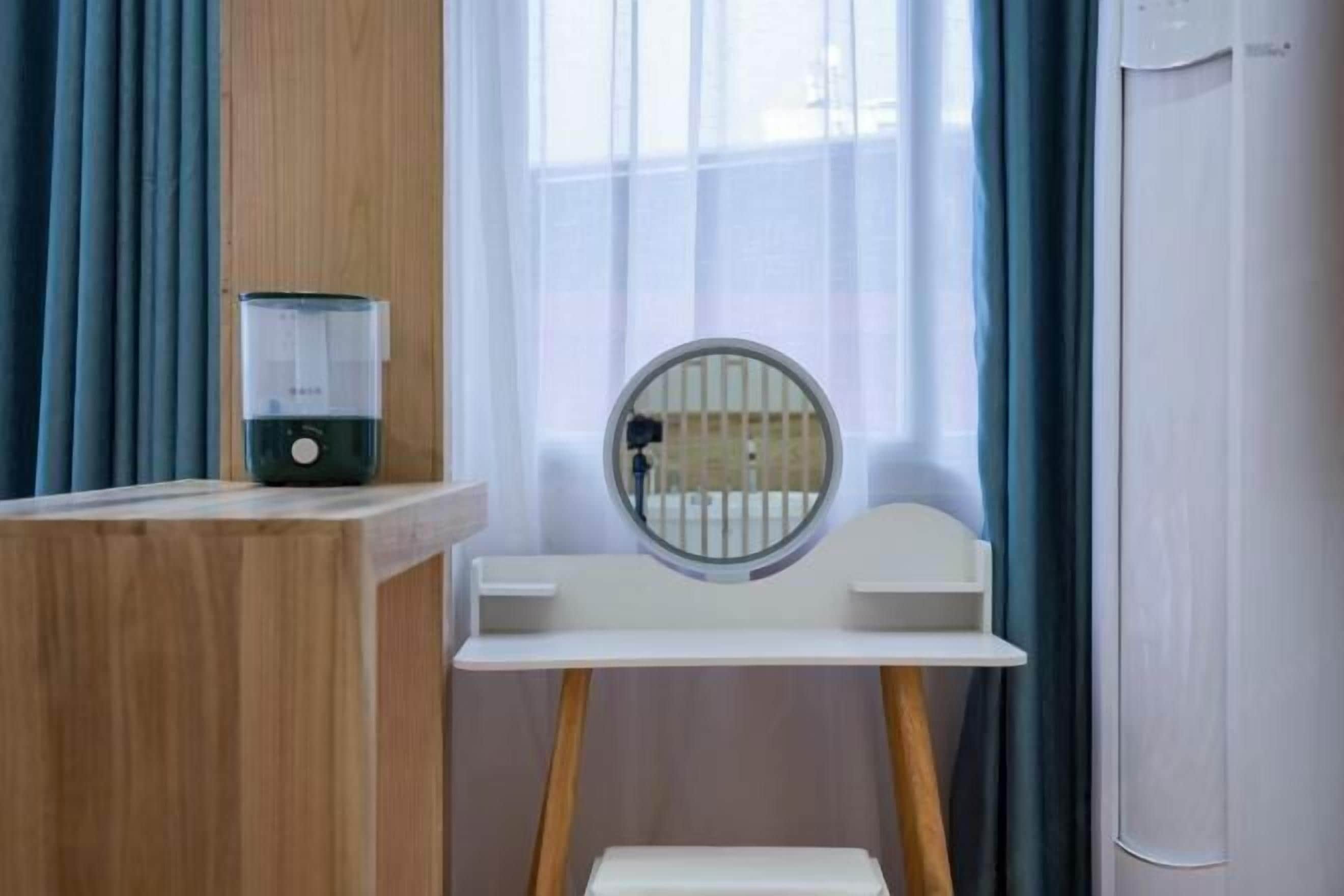 Quarto com escrivaninha de madeira, um espelho redondo pequeno, cadeira branca e janela com cortinas.