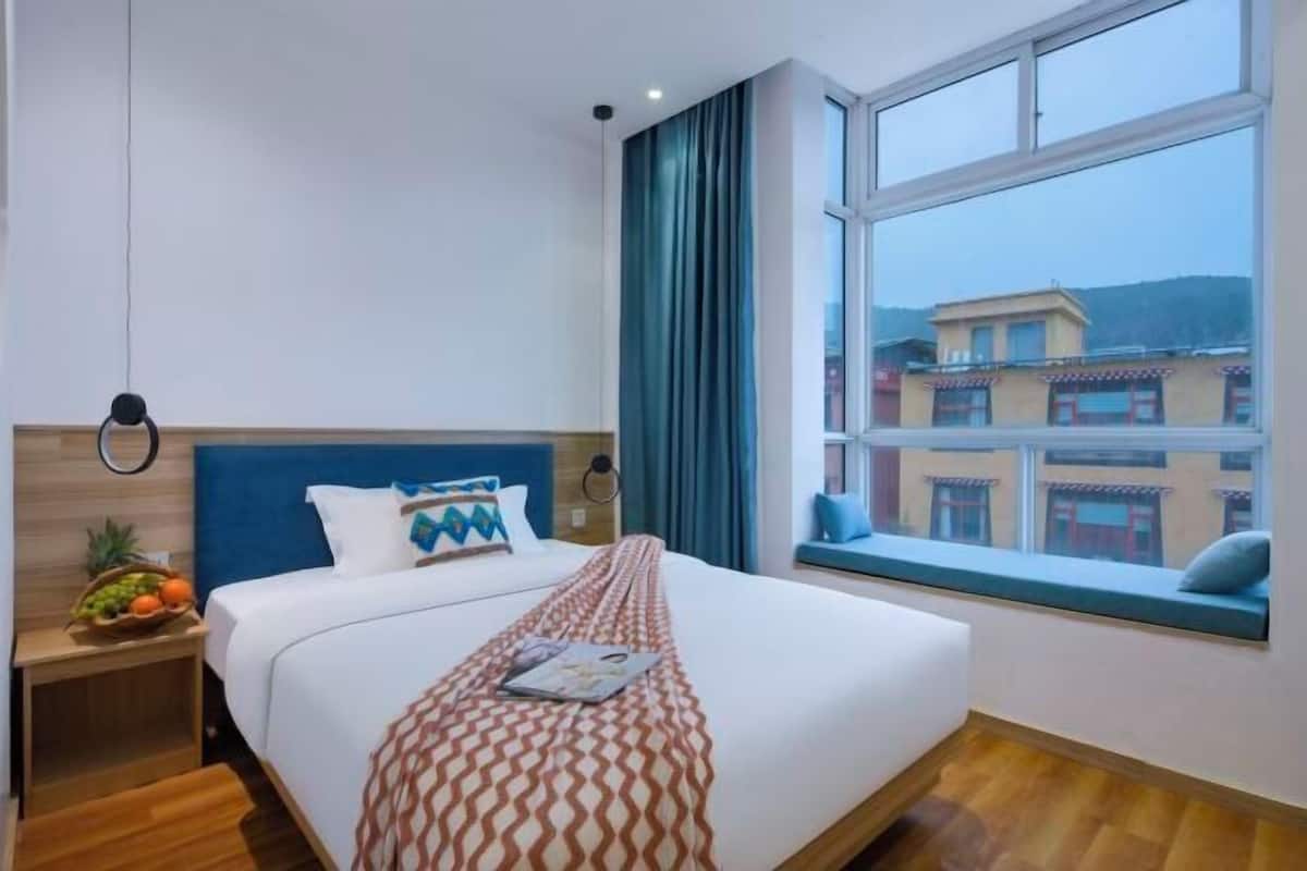 Une chambre d’hôtel avec un grand lit, une fenêtre donnant sur des immeubles, une table de chevet avec des fruits et une tête de lit bleue.