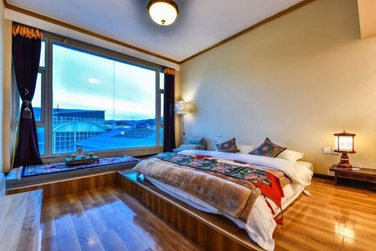 Une chambre avec une grande fenêtre, un lit avec une couverture, une table de chevet avec une lampe, et une vue sur un pont.