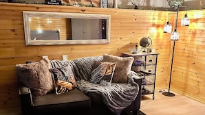 Une pièce chaleureuse avec un mur en bois, un grand miroir, un canapé gris agrémenté de coussins décoratifs, une petite table d’appoint et un lampadaire.