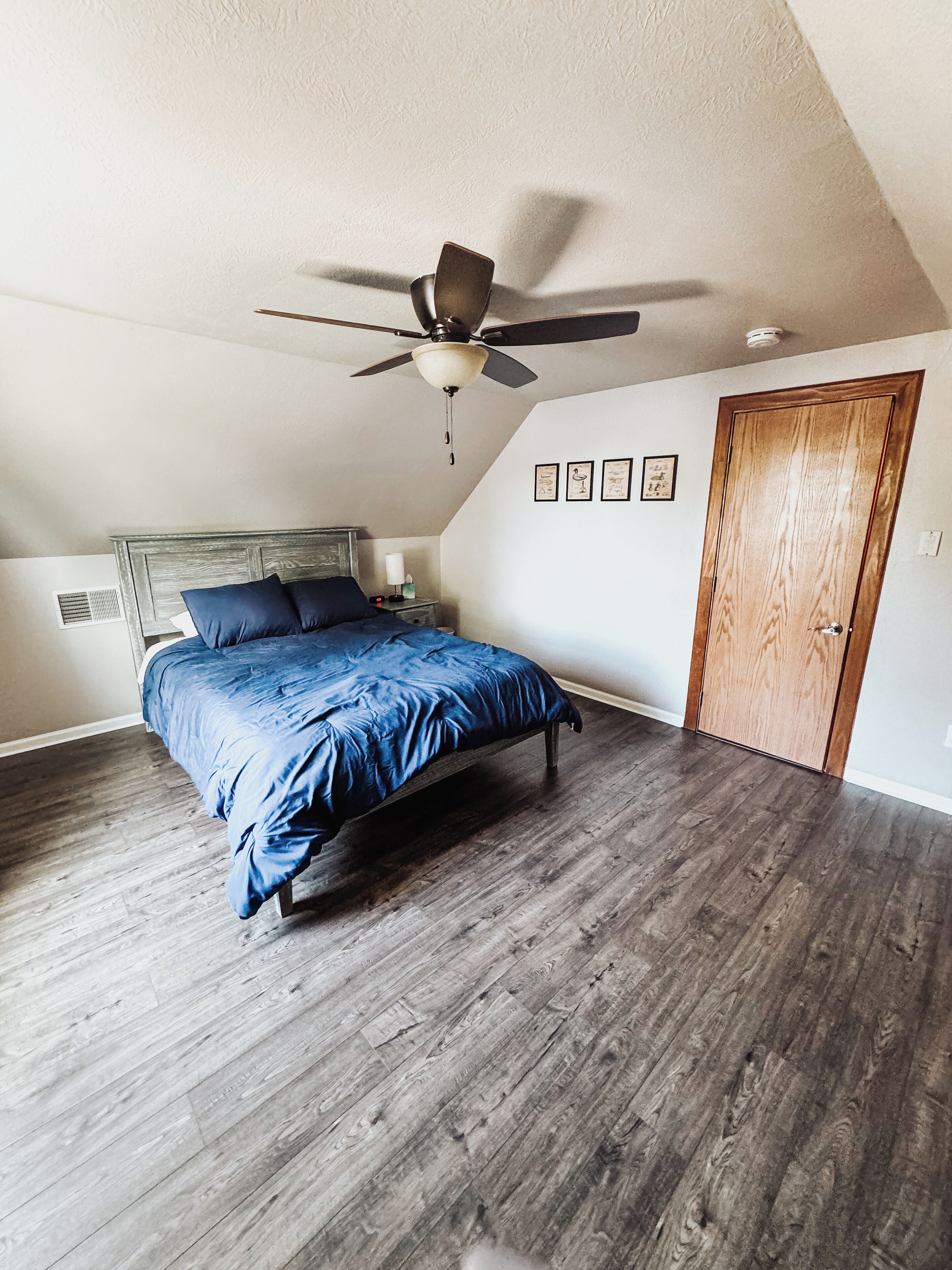 A bedroom with a bed, wooden flooring, a ceiling fan, and a wooden door.