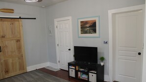 Una habitación con una puerta de madera, una puerta blanca, un televisor sobre un soporte negro y un cuadro en la pared.