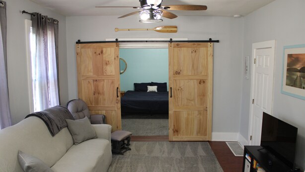 Una habitación con sofá, televisión, ventilador de techo y una puerta corrediza que da a un dormitorio con cama y mesita de noche.
