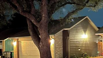 A residential property with a large tree in front of a garage, a well-lit driveway, and a house with a sloped roof.